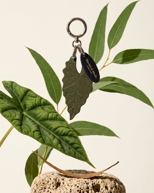 A Maverick Made. leather leaf keychain charm from Singapore, photographed suspended above porous natural stone and layered tropical foliage. The handcrafted olive green leather charm features engraved leaf vein detailing, polished silver-tone hardware and a branded leather tag. Soft natural lighting, botanical styling and a clean neutral backdrop create a refined editorial image for artisan leather accessories, handcrafted bag charms and unique leather gifts in Singapore.