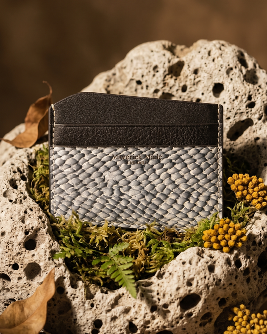 A handcrafted Maverick Made. leather cardholder from Singapore, made with grey textured salmon skin leather and black full-grain leather. The slim cardholder is photographed upright on porous white stone, surrounded by green moss, dried brown leaves, grey rocks and small yellow wildflowers. Warm directional lighting, shallow depth of field and earthy styling create a rugged nature-inspired product image for premium leather goods, artisan wallets and unique cardholders in Singapore.