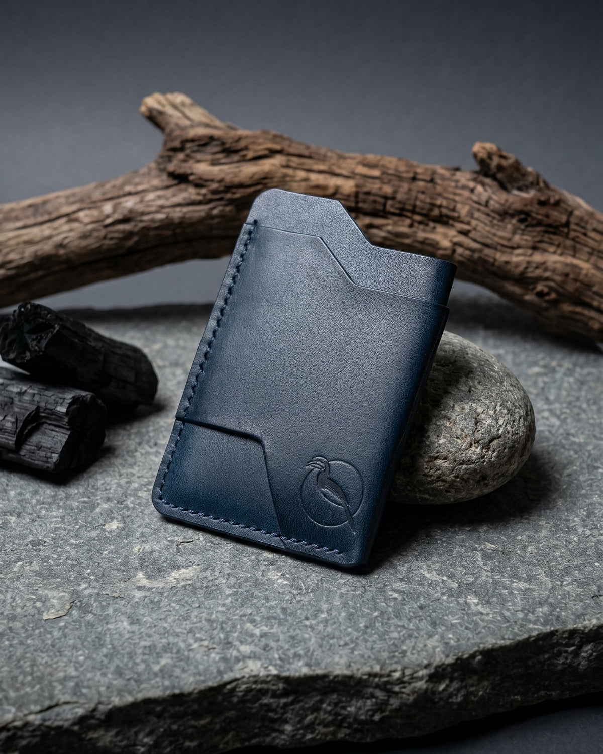 Handcrafted indigo blue leather card holder by Maverick Made. in Singapore, featuring hand stitching, an embossed hornbill logo, and a slim minimalist wallet design photographed on stone, driftwood and charcoal.
