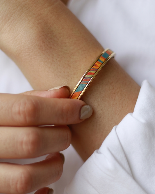 A Maverick Made. PVD chrome plated stainless steel leather cuff bracelet from Singapore, featuring a colourful striped leather inlay with warm orange, blue and red tones. The bracelet is worn with a white shirt in a soft minimalist portrait, with the wearer gently touching the cuff. Natural lighting, neutral styling and shallow depth of field create a refined editorial image for premium leather jewellery, artisan bracelets and handcrafted fashion accessories in Singapore.