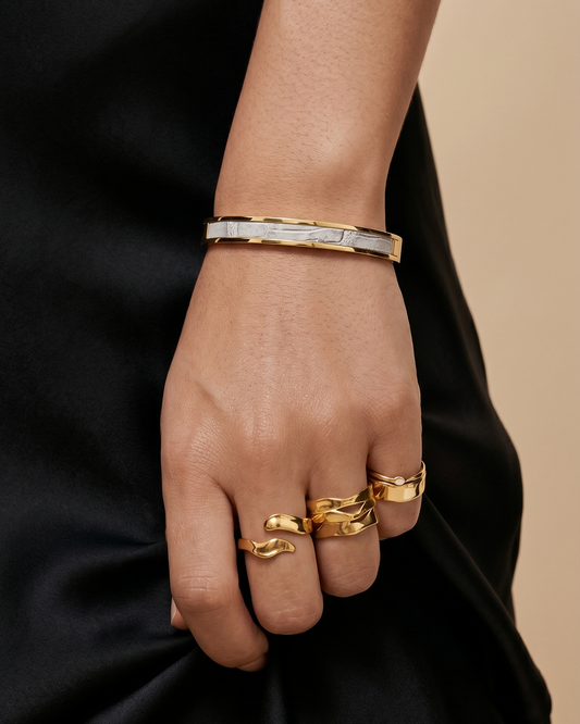 A Maverick Made. gold stainless steel leather cuff bracelet from Singapore, worn with sculptural gold rings and a black satin outfit. The slim cuff features polished gold-tone metal with a leather inlay, photographed in soft warm lighting against a neutral backdrop. Clean composition, refined styling and shallow depth of field create a luxury editorial image for premium leather jewellery, artisan bracelets and handcrafted fashion accessories in Singapore.