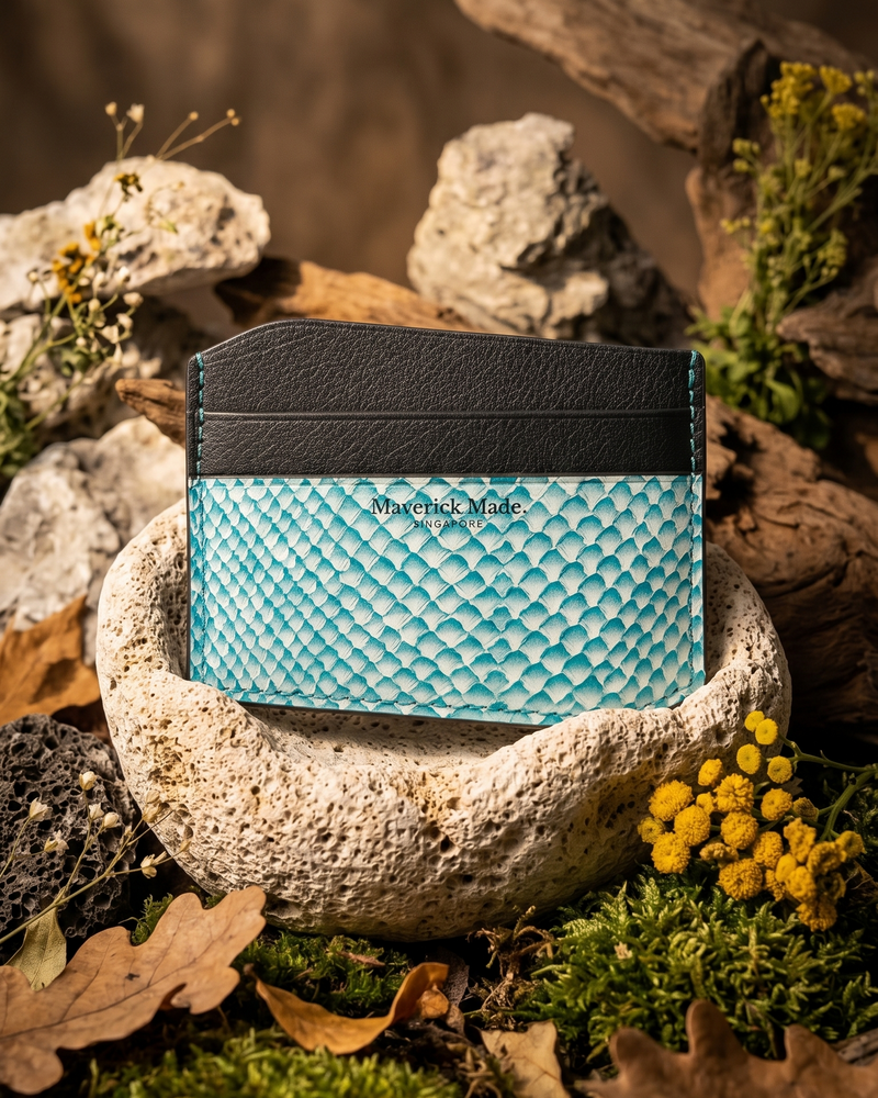 A handcrafted Maverick Made. leather cardholder from Singapore, made with turquoise textured salmon skin leather and black full-grain leather, photographed in a rugged nature-inspired display. The cardholder sits upright inside a porous stone bowl, surrounded by moss, dried leaves, driftwood, rocks and small yellow flowers. Warm directional lighting, earthy tones and shallow depth of field create an artisanal, outdoor-inspired product image for premium leather goods.