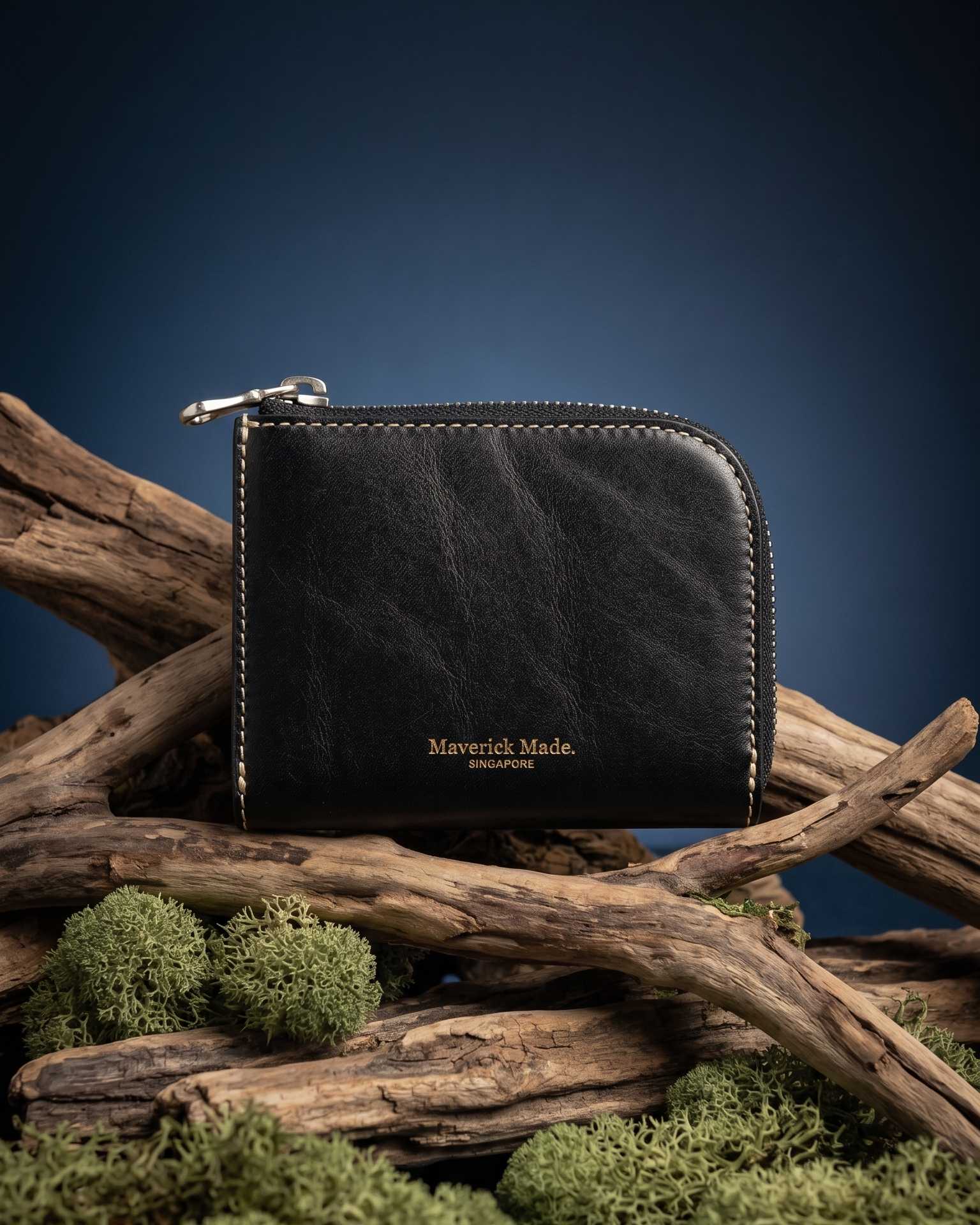 A black leather zip wallet by Maverick Made. Singapore, styled upright on layered driftwood with pale green moss and lichen in the foreground. The compact wallet features a rounded corner, visible zipper pull, contrast stitching and gold Maverick Made. Singapore logo stamping. A dark midnight blue gradient background and soft directional studio lighting create a rugged yet refined product photograph for premium leather goods, handcrafted wallets, artisan leather accessories and luxury everyday carry in Sing