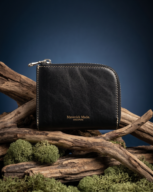 A black leather zip wallet by Maverick Made. Singapore, styled upright on layered driftwood with pale green moss and lichen in the foreground. The compact wallet features a rounded corner, visible zipper pull, contrast stitching and gold Maverick Made. Singapore logo stamping. A dark midnight blue gradient background and soft directional studio lighting create a rugged yet refined product photograph for premium leather goods, handcrafted wallets, artisan leather accessories and luxury everyday carry in Sing
