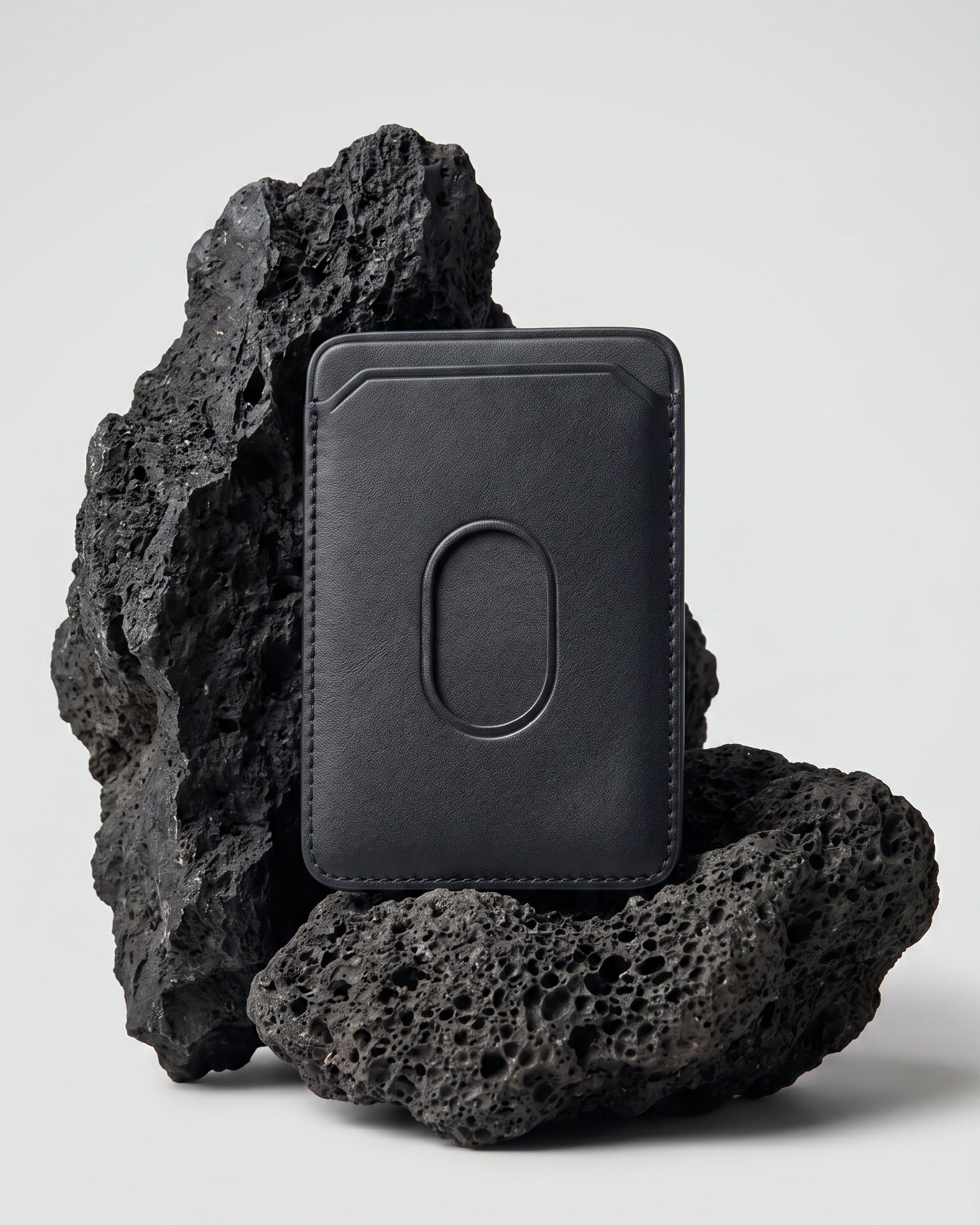 A black Maverick Made. leather MagSafe cardholder from Singapore, styled upright among porous black volcanic rocks. The minimalist cardholder features smooth black leather, rounded corners, stitched edges, angled card slot details and an oval MagSafe impression on the back. Soft studio lighting and a clean grey background create a refined product image for premium leather accessories, handcrafted cardholders, minimalist wallets and everyday carry in Singapore.