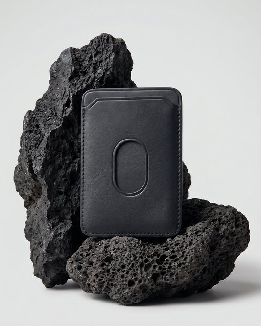 A black Maverick Made. leather MagSafe cardholder from Singapore, styled upright among porous black volcanic rocks. The minimalist cardholder features smooth black leather, rounded corners, stitched edges, angled card slot details and an oval MagSafe impression on the back. Soft studio lighting and a clean grey background create a refined product image for premium leather accessories, handcrafted cardholders, minimalist wallets and everyday carry in Singapore.