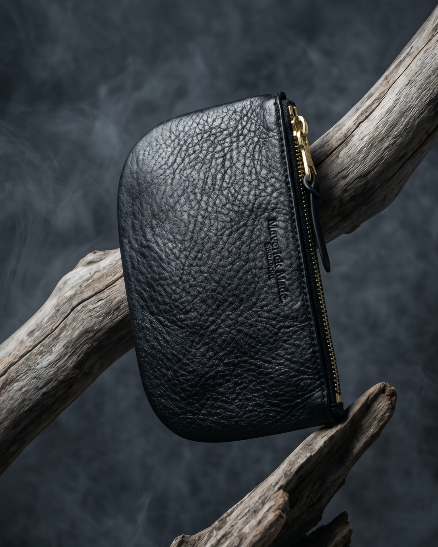 A black leather zipper pouch by Maverick Made. Singapore, suspended among sculptural driftwood against a dark smoky backdrop. The pouch features textured full-grain leather, a curved compact silhouette, brass zipper, leather pull tab and subtle embossed branding. Directional lighting, smoke and weathered wood create a rugged editorial image for premium leather goods, artisan zipper pouches and handcrafted everyday carry in Singapore.