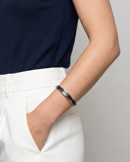 A Maverick Made. blue leather cuff bracelet from Singapore, featuring polished gold-tone stainless steel with a deep blue leather inlay. The bracelet is worn with a navy sleeveless knit top and tailored white trousers in a clean minimalist studio portrait. Soft lighting, neutral backdrop and refined styling create an elegant editorial image for premium leather jewellery, artisan bracelets and handcrafted fashion accessories in Singapore.