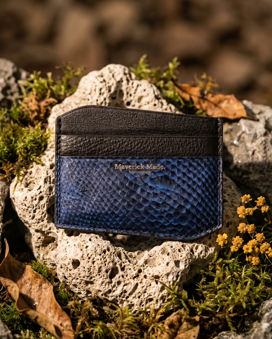 A handcrafted Maverick Made. leather cardholder from Singapore, made with navy blue textured salmon skin leather and black full-grain leather. The slim cardholder is photographed upright on porous white stone, surrounded by green moss, dried brown leaves, grey rocks and small yellow wildflowers. Warm directional lighting, shallow depth of field and earthy styling create a rugged nature-inspired product image for premium leather goods, artisan wallets and unique cardholders in Singapore.