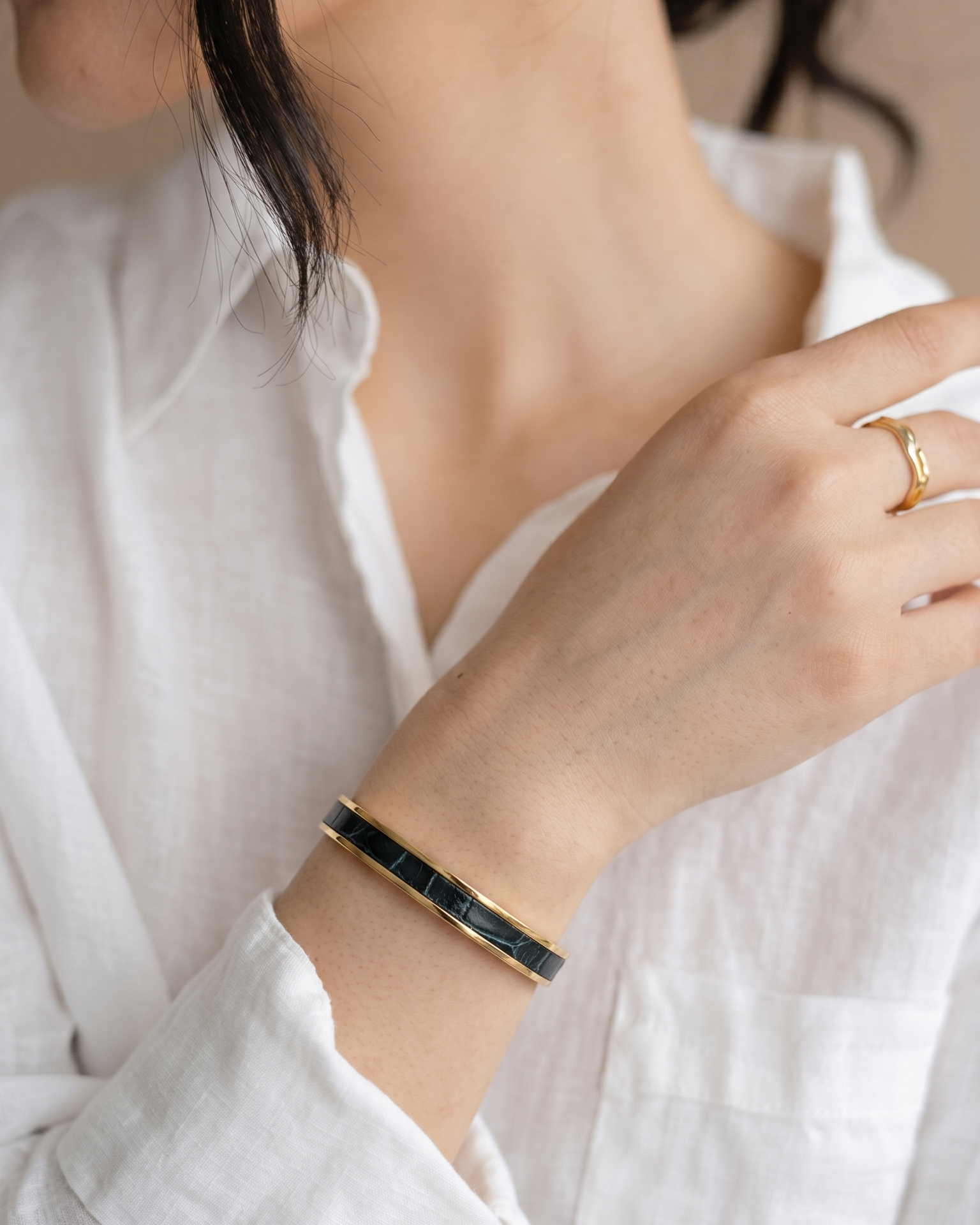 A Maverick Made. gold stainless steel leather cuff bracelet from Singapore, featuring a dark leather inlay and polished gold-tone finish. The bracelet is worn with a white linen shirt, gold ring and hoop earrings in a soft natural light portrait. Neutral tones, shallow depth of field and clean styling create an elegant editorial image for premium leather jewellery, artisan bracelets and handcrafted fashion accessories in Singapore.