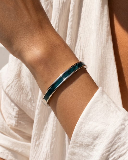 A Maverick Made. stainless steel leather cuff bracelet from Singapore, featuring a deep teal leather inlay and polished silver-tone finish. The bracelet is worn with a lightweight white linen outfit under warm natural sunlight, creating soft highlights and natural skin tones. Minimal styling, shallow depth of field and clean composition create a refined editorial image for premium leather jewellery, artisan bracelets and handcrafted fashion accessories in Singapore.