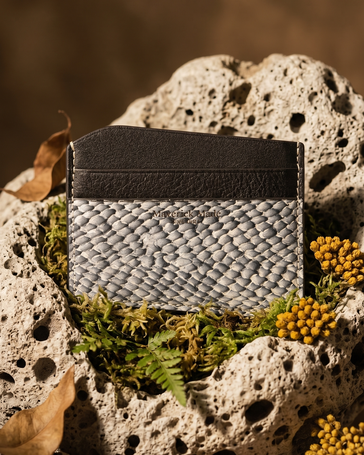 A handcrafted Maverick Made. leather cardholder from Singapore, made with grey textured salmon skin leather and black full-grain leather. The slim cardholder is photographed upright on porous white stone, surrounded by green moss, dried brown leaves, grey rocks and small yellow wildflowers. Warm directional lighting, shallow depth of field and earthy styling create a rugged nature-inspired product image for premium leather goods, artisan wallets and unique cardholders in Singapore.