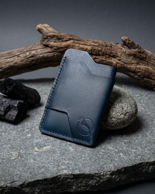 Handcrafted indigo blue leather card holder by Maverick Made. in Singapore, featuring hand stitching, an embossed hornbill logo, and a slim minimalist wallet design photographed on stone, driftwood and charcoal.