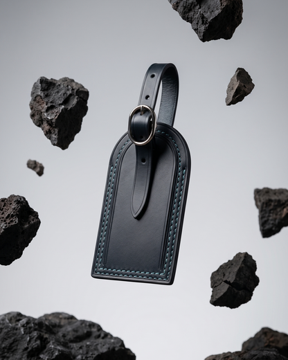 An indigo blue leather luggage tag by Maverick Made. Singapore, photographed floating above a gravel ground plane and surrounded by suspended porous volcanic rocks. The tag features a rounded top, stitched leather edges, raised border detailing, adjustable leather strap and polished metal ring. Soft grey studio background, directional lighting and sharp product focus create a modern editorial image for premium leather travel accessories, personalised luggage tags and handcrafted leather goods in Singapore.