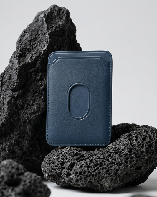 An indigo blue Maverick Made. leather MagSafe cardholder from Singapore, styled upright among porous black volcanic rocks. The minimalist cardholder features smooth black leather, rounded corners, stitched edges, angled card slot details and an oval MagSafe impression on the back. Soft studio lighting and a clean grey background create a refined product image for premium leather accessories, handcrafted cardholders, minimalist wallets and everyday carry in Singapore.