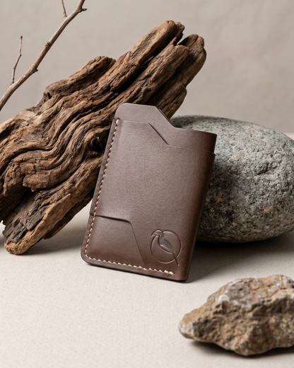 A handcrafted brown leather cardholder by Maverick Made. Singapore, photographed upright beside weathered driftwood, smooth stone and natural textures. The slim minimalist wallet features clean geometric card slots, visible stitching and subtle embossed artwork detail on premium leather. Soft natural lighting, earthy tones and organic styling create a refined editorial image for artisan leather goods, handcrafted cardholders and premium everyday carry accessories in Singapore.