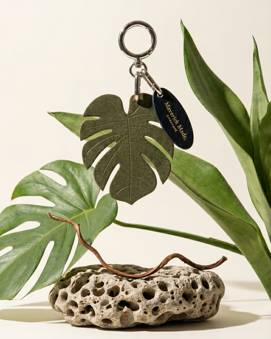 A Maverick Made. leather monstera leaf keychain charm from Singapore, photographed suspended above porous natural stone and surrounded by lush tropical foliage. The handcrafted green leather charm features detailed leaf cut-outs, polished metal hardware and a branded leather tag. Soft natural lighting, neutral backdrop and botanical styling create a clean editorial image for artisan leather accessories, handcrafted bag charms and unique leather gifts in Singapore.