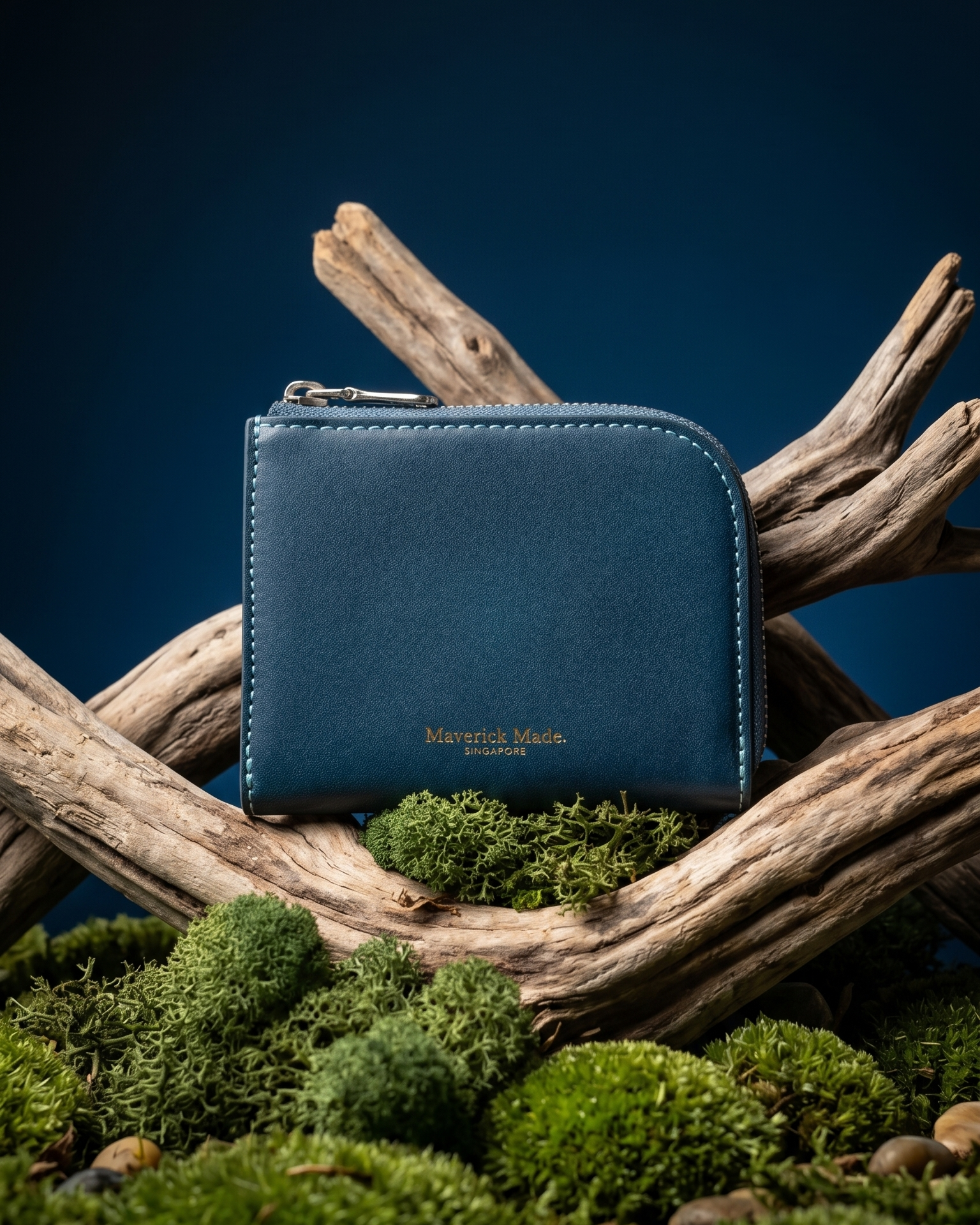 A blue leather zip wallet by Maverick Made. Singapore, styled upright on layered driftwood with pale green moss and lichen in the foreground. The compact wallet features a rounded corner, visible zipper pull, contrast stitching and gold Maverick Made. Singapore logo stamping. A dark midnight blue gradient background and soft directional studio lighting create a rugged yet refined product photograph for premium leather goods, handcrafted wallets, artisan leather accessories and luxury carry in Singapore.
