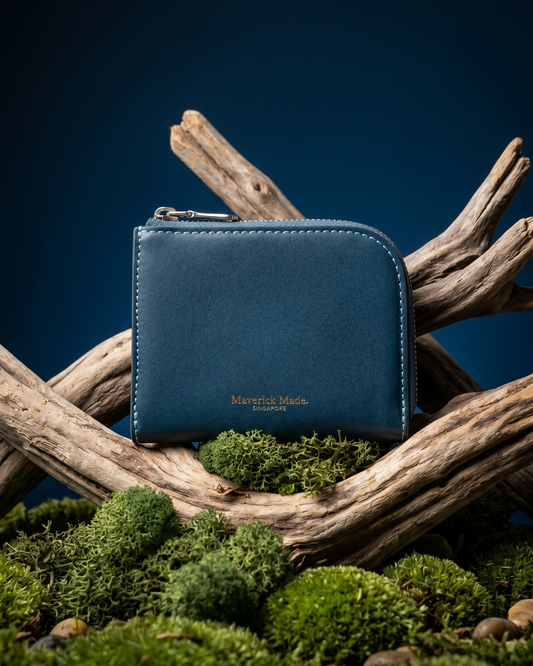 A blue leather zip wallet by Maverick Made. Singapore, styled upright on layered driftwood with pale green moss and lichen in the foreground. The compact wallet features a rounded corner, visible zipper pull, contrast stitching and gold Maverick Made. Singapore logo stamping. A dark midnight blue gradient background and soft directional studio lighting create a rugged yet refined product photograph for premium leather goods, handcrafted wallets, artisan leather accessories and luxury carry in Singapore.