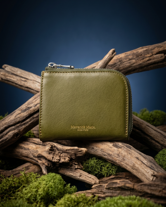 A green leather zip wallet by Maverick Made. Singapore, styled upright on layered driftwood with pale green moss and lichen in the foreground. The compact wallet features a rounded corner, visible zipper pull, contrast stitching and gold Maverick Made. Singapore logo stamping. A dark midnight blue gradient background and soft directional studio lighting create a rugged yet refined product photograph for premium leather goods, handcrafted wallets, artisan leather accessories and luxury carry in Singapore
