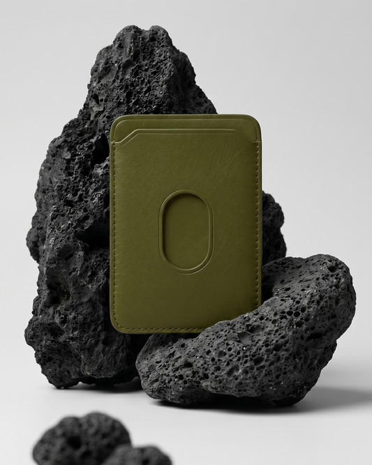 An olive green Maverick Made. leather MagSafe cardholder from Singapore, styled upright among porous black volcanic rocks. The minimalist cardholder features smooth black leather, rounded corners, stitched edges, angled card slot details and an oval MagSafe impression on the back. Soft studio lighting and a clean grey background create a refined product image for premium leather accessories, handcrafted cardholders, minimalist wallets and everyday carry in Singapore.