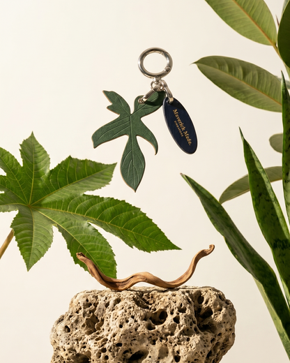 A Maverick Made. leather leaf keychain charm from Singapore, photographed suspended above porous natural stone and surrounded by tropical green plants. The handcrafted green leather charm features engraved leaf detailing, polished silver-tone hardware and a branded leather tag. Soft natural lighting, botanical styling and a clean neutral backdrop create a refined editorial image for artisan leather accessories, handcrafted bag charms and unique leather gifts in Singapore.