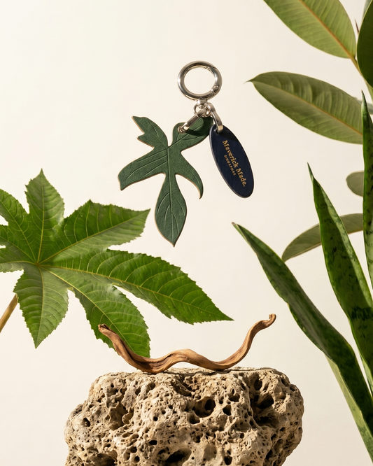 A Maverick Made. leather leaf keychain charm from Singapore, photographed suspended above porous natural stone and surrounded by tropical green plants. The handcrafted green leather charm features engraved leaf detailing, polished silver-tone hardware and a branded leather tag. Soft natural lighting, botanical styling and a clean neutral backdrop create a refined editorial image for artisan leather accessories, handcrafted bag charms and unique leather gifts in Singapore.