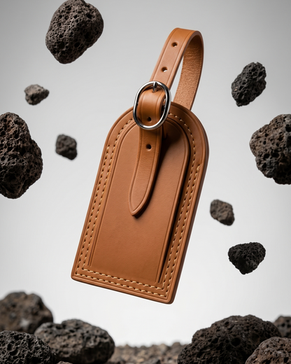 A tan leather luggage tag by Maverick Made. Singapore, photographed floating above a gravel ground plane and surrounded by suspended porous volcanic rocks. The tag features a rounded top, stitched leather edges, raised border detailing, adjustable leather strap and polished metal ring. Soft grey studio background, directional lighting and sharp product focus create a modern editorial image for premium leather travel accessories, personalised luggage tags and handcrafted leather goods in Singapore.
