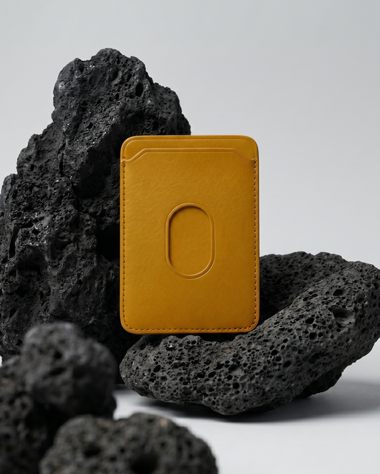 A tan Maverick Made. leather MagSafe cardholder from Singapore, styled upright among porous black volcanic rocks. The minimalist cardholder features smooth black leather, rounded corners, stitched edges, angled card slot details and an oval MagSafe impression on the back. Soft studio lighting and a clean grey background create a refined product image for premium leather accessories, handcrafted cardholders, minimalist wallets and everyday carry in Singapore.