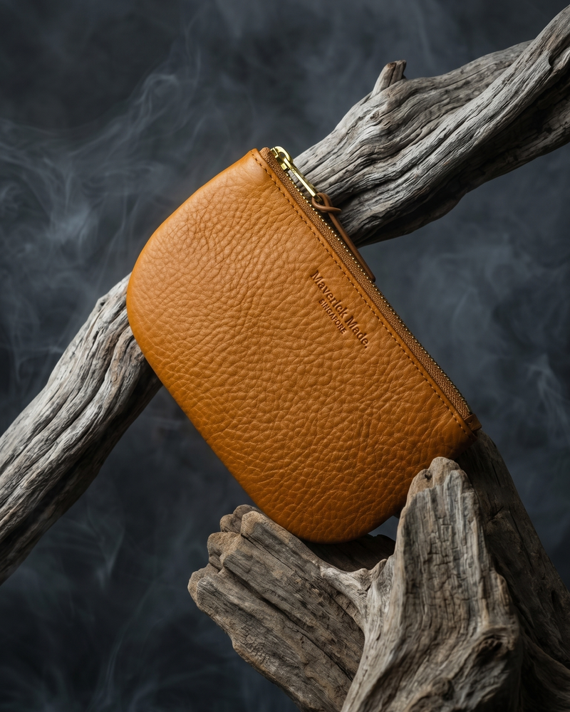 A tan leather zipper pouch by Maverick Made. Singapore, suspended among sculptural driftwood against a dark smoky backdrop. The pouch features textured full-grain leather, a curved compact silhouette, brass zipper, leather pull tab and subtle embossed branding. Directional lighting, smoke and weathered wood create a rugged editorial image for premium leather goods, artisan zipper pouches and handcrafted everyday carry in Singapore.