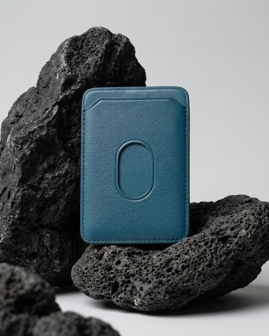 A blue Maverick Made. leather MagSafe cardholder from Singapore, styled upright among porous black volcanic rocks. The minimalist cardholder features smooth black leather, rounded corners, stitched edges, angled card slot details and an oval MagSafe impression on the back. Soft studio lighting and a clean grey background create a refined product image for premium leather accessories, handcrafted cardholders, minimalist wallets and everyday carry in Singapore.