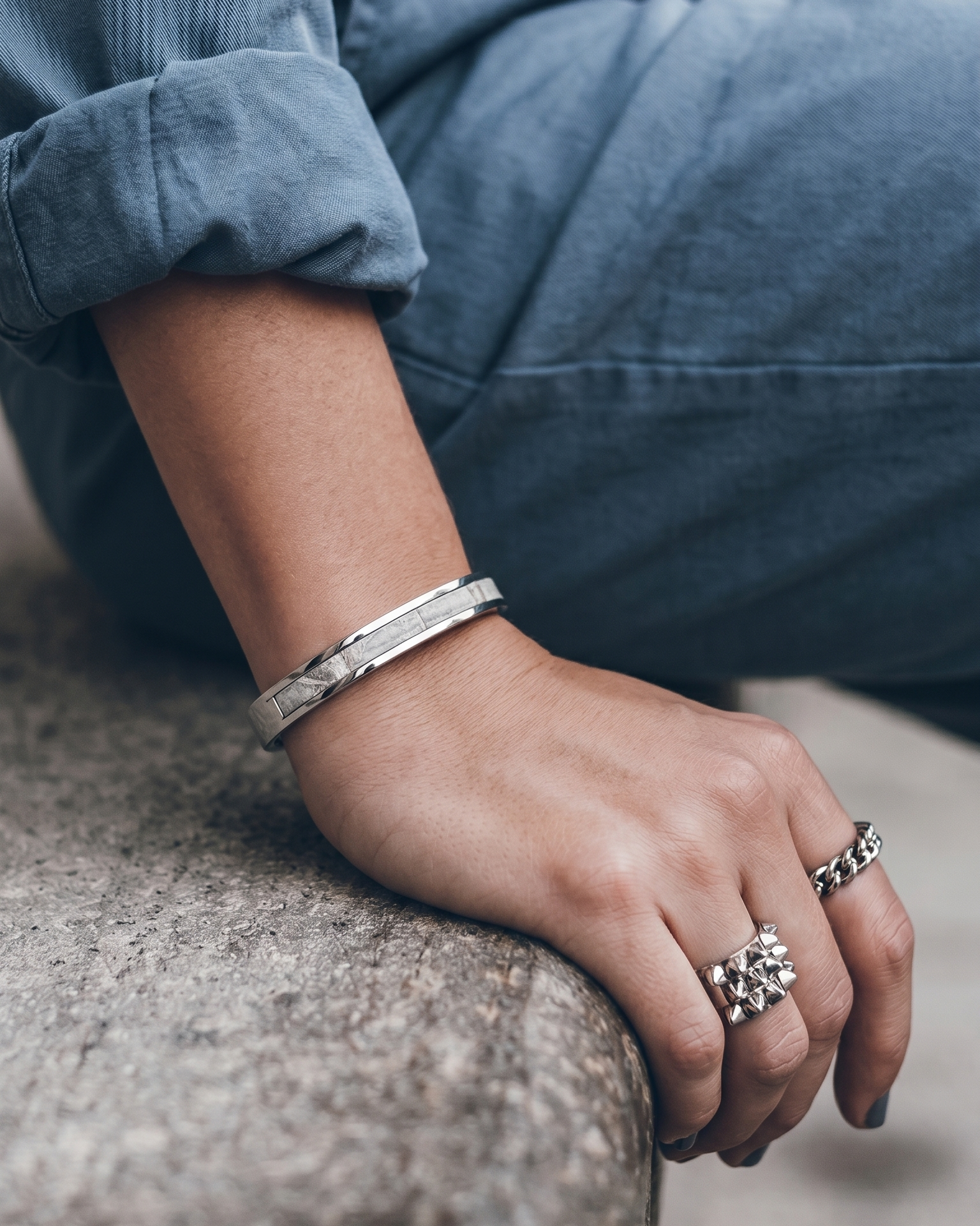 A Maverick Made. stainless steel leather cuff bracelet from Singapore, photographed on a seated person wearing rolled denim sleeves and silver rings. The slim cuff features polished metal construction with a leather inlay, styled in a clean urban lifestyle setting with soft natural light and shallow depth of field. Minimalist composition and muted tones create a refined editorial image for premium leather jewellery, artisan accessories and handcrafted fashion pieces in Singapore.
