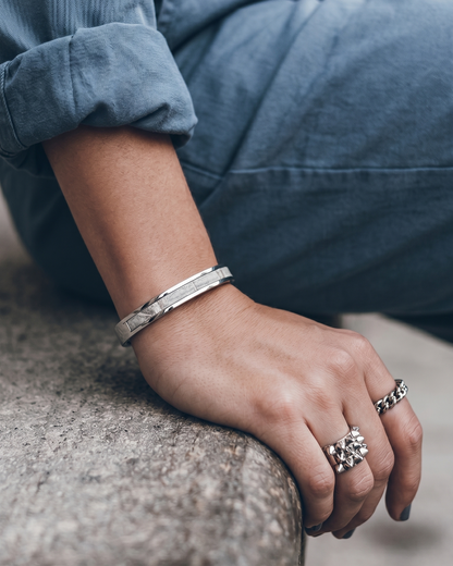 A Maverick Made. stainless steel leather cuff bracelet from Singapore, photographed on a seated person wearing rolled denim sleeves and silver rings. The slim cuff features polished metal construction with a leather inlay, styled in a clean urban lifestyle setting with soft natural light and shallow depth of field. Minimalist composition and muted tones create a refined editorial image for premium leather jewellery, artisan accessories and handcrafted fashion pieces in Singapore.